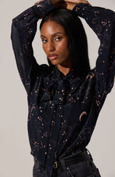 A woman is wearing a long-sleeved, dark satin blouse with a whimsical celestial print featuring stars, clouds, and crescent moons in light-colored shades. The blouse has a large bow tied at the collar. She pairs it with black jeans and a matching black belt.